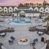  A fire pit is surrounded by chairs and tables in a courtyard with a pool behind it.
